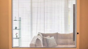 A room with a window with horizontal blinds, a bed, and a small shelf with items on it.