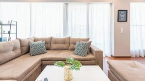 A living room with a beige sectional sofa, patterned throw pillows, a coffee table with a plant, and large windows with sheer curtains.