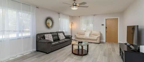 Un salon avec un canapé gris, un canapé beige, une table basse en verre, un ventilateur de plafond et un téléviseur posé sur un meuble noir.