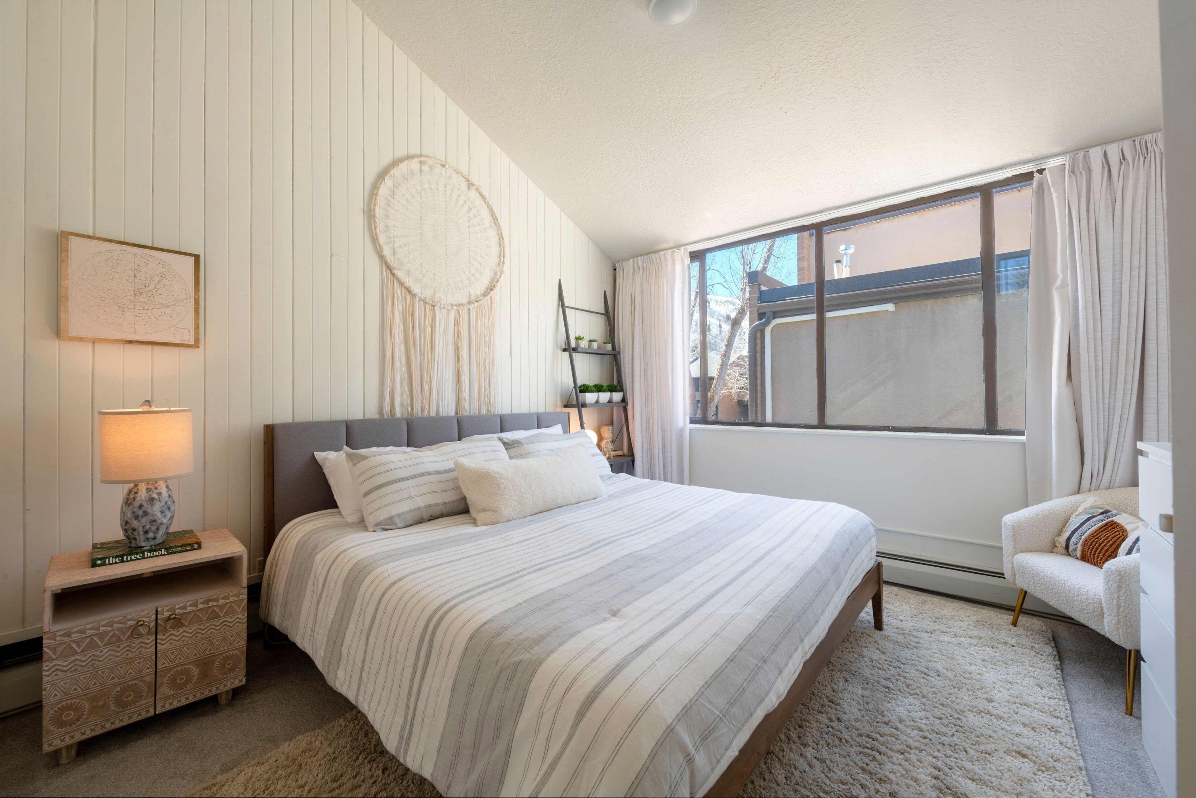 A bedroom with a large bed, a nightstand, a chair, a window with curtains, and a wall-mounted shelf.
