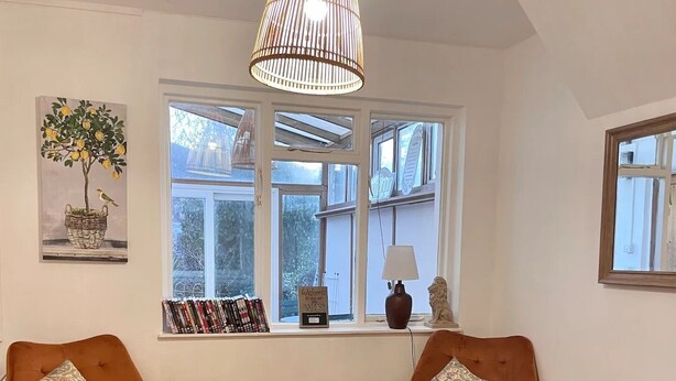 Une pièce chaleureuse avec deux fauteuils orange, une table basse en bois, un luminaire suspendu, une bibliothèque remplie de livres et une fenêtre donnant sur des arbres.
