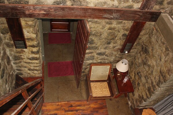 A narrow stone-walled stairway with a wooden handrail and red carpeted steps leading to a room with a wooden chair, a small table with a lamp, and a wicker basket.