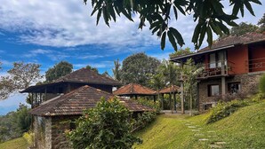 A hillside property with multiple buildings, including a stone house and a red building with a balcony. - Misty Hogans Hill Station Stay | Entire Stay Near Munnar | Sleeps 10 (Kambilikandam)