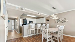 A modern kitchen with white cabinets, a central island, and a dining area with wooden chairs and a table. - Sunrise Pointe 9E-Oceanfront 3BR-Pool, Hot Tub (North Myrtle Beach)
