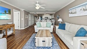 A spacious living room with a wooden coffee table, a white sofa with blue cushions, a dining area with white chairs, and a kitchen in the background. - Sunrise Pointe 9E-Oceanfront 3BR-Pool, Hot Tub (North Myrtle Beach)