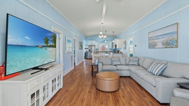 A spacious living room with a large flat-screen TV, a grey sectional sofa, a round ottoman, and a kitchen in the background. - The Reagan- 4BR Channel House-Hot Tub (North Myrtle Beach)
