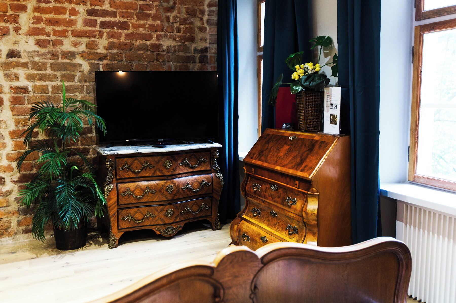 Ein Zimmer mit Backsteinwand, einer Holzkommode mit Schubladen, einem Fernseher auf einem Standfuß und einem Fenster mit Vorhängen.