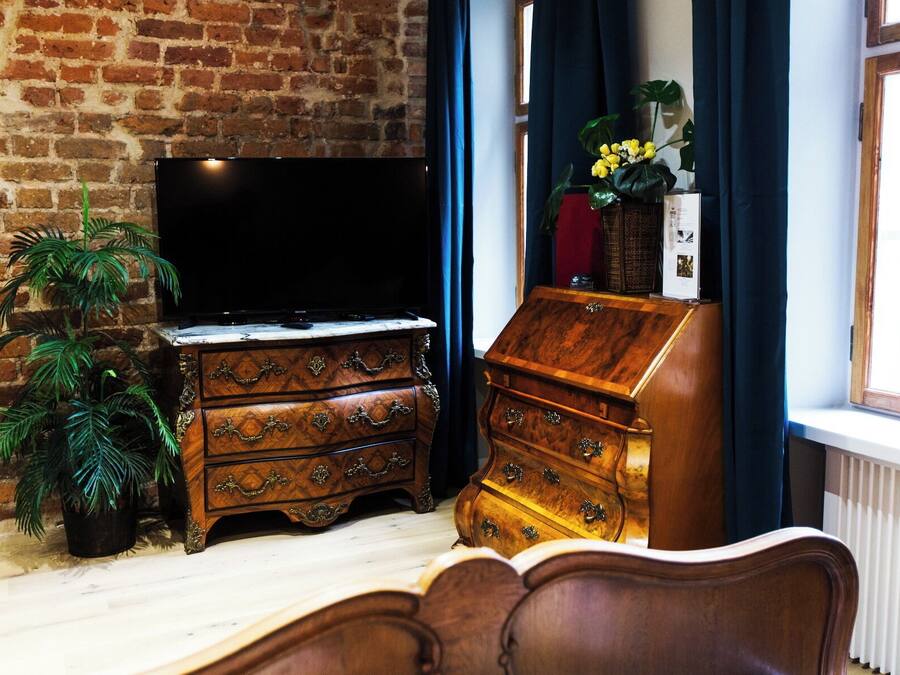 Ein Zimmer mit Backsteinwand, einer Holzkommode mit Schubladen, einem Fernseher auf einem Standfuß und einem Fenster mit Vorhängen.