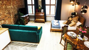 Una sala de estar acogedora con pared de ladrillo, sofá verde, cómoda de madera, televisor y cama con almohadas y manta.