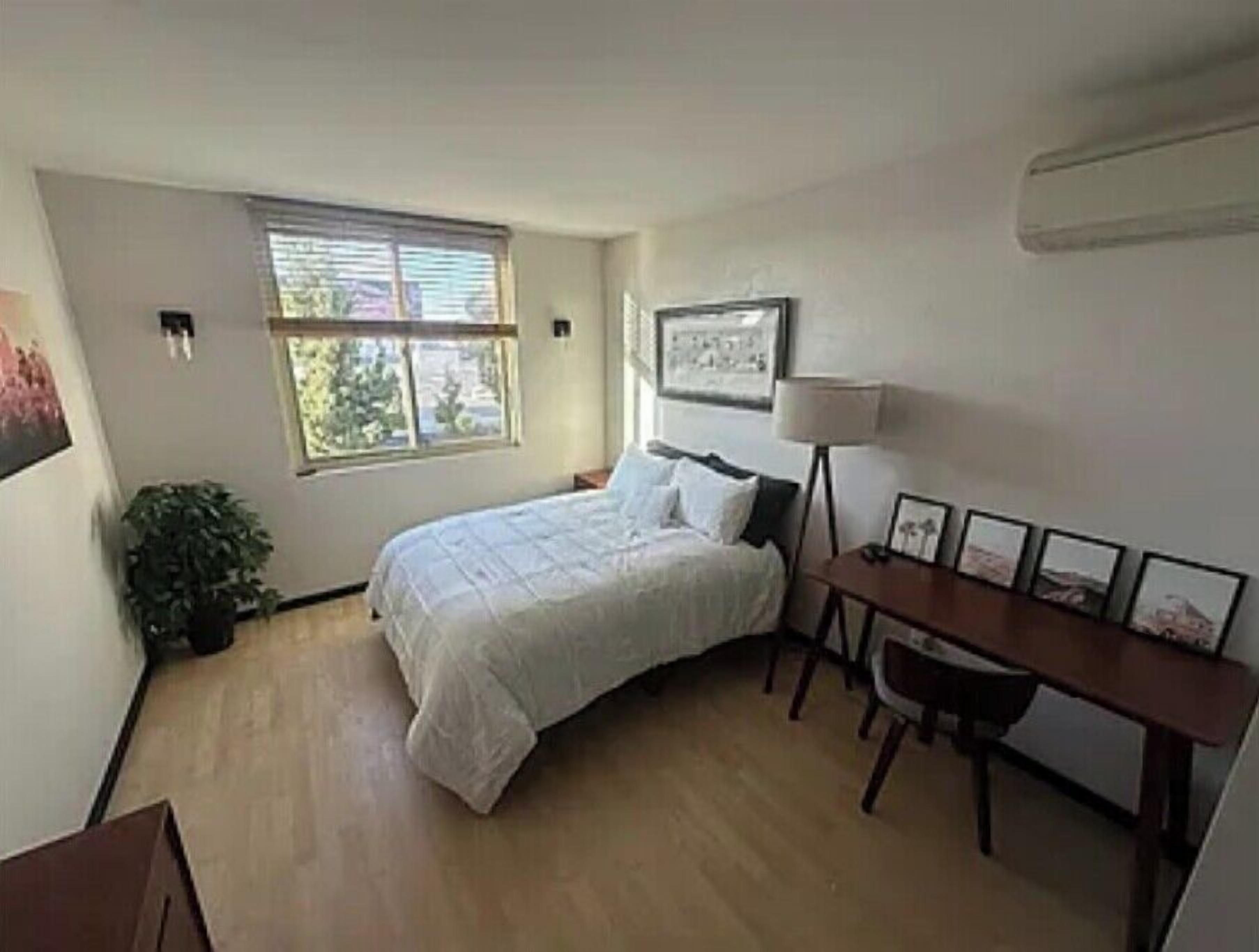 A bedroom with a bed, a desk, a chair, a window, and a plant.