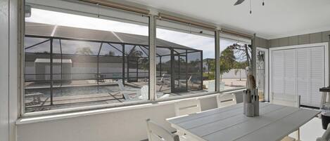 A room with a large window overlooking a pool area, a ceiling fan, and a table with chairs.