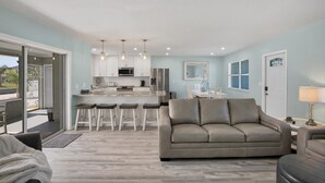Ein modernes Wohnzimmer mit grauem Sofa, einem Essbereich mit Barhocker, einer Küche mit weißen Schränken und Edelstahlgeräten sowie einer Glastür zum Balkon.