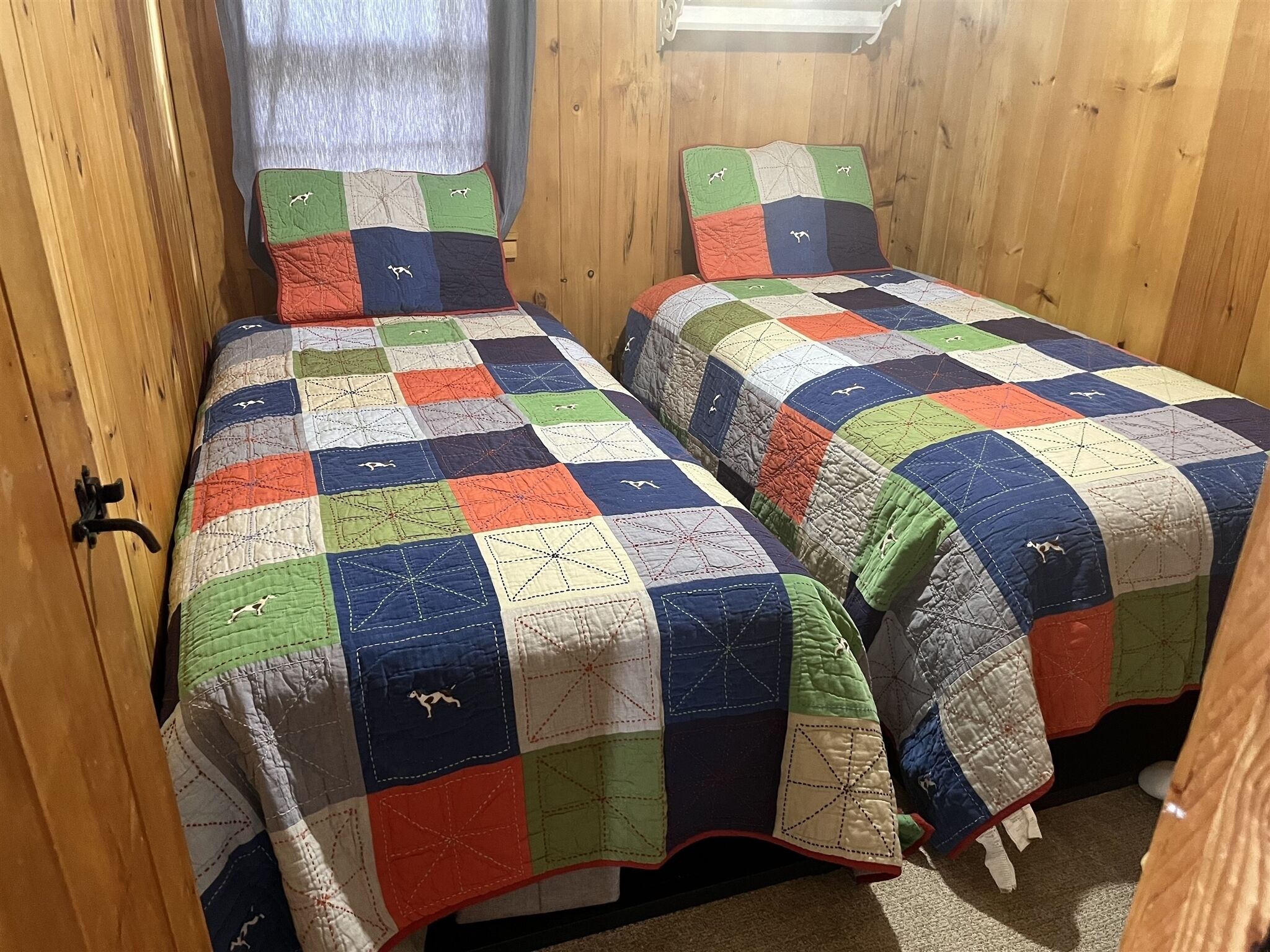 Dos camas individuales con colchas coloridas, paredes de madera y una ventana con cortinas.