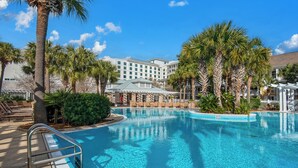 A hotel pool area with a clear blue pool, palm trees, lounge chairs, and a building with multiple windows in the background.