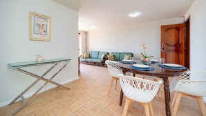 A dining area with a glass-top table, white chairs, a wooden dining table, and a green sofa in the background. - Cf05 Prédio beira mar Apto 3 quartos (Cabo Frio)