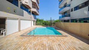 A swimming pool area with a tiled deck, surrounded by multi-story buildings with balconies. - Cf05 Prédio beira mar Apto 3 quartos (Cabo Frio)
