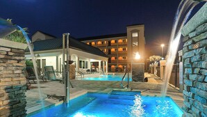 Un área de piscina iluminada durante la noche, con un edificio al fondo.