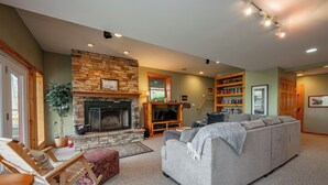 A cozy living room with a stone fireplace, a grey sofa, a wooden rocking chair, and a bookshelf. - Chic Dog Friendly Chalet w/ Fire Pit & Hot Tub (McHenry)