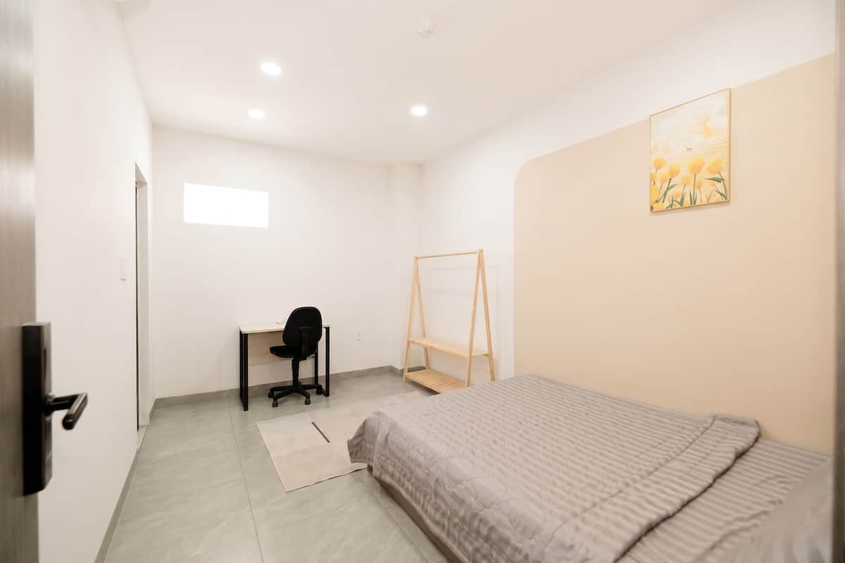 A bedroom with a bed, a desk, a chair, a hanging shelf, and a painting on the wall.