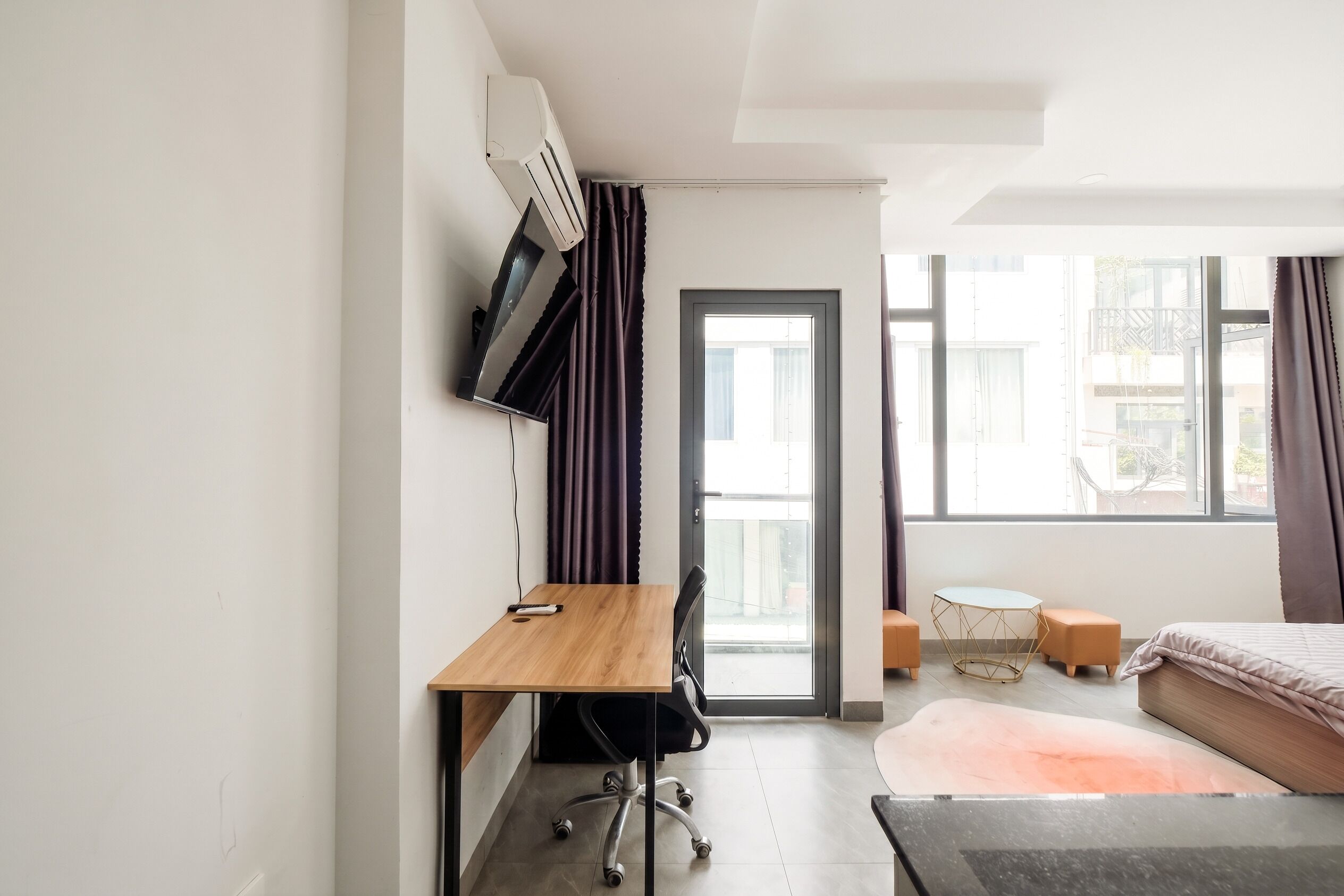 A modern hotel room with a wooden desk, a bed, a small table, and a TV mounted on the wall.