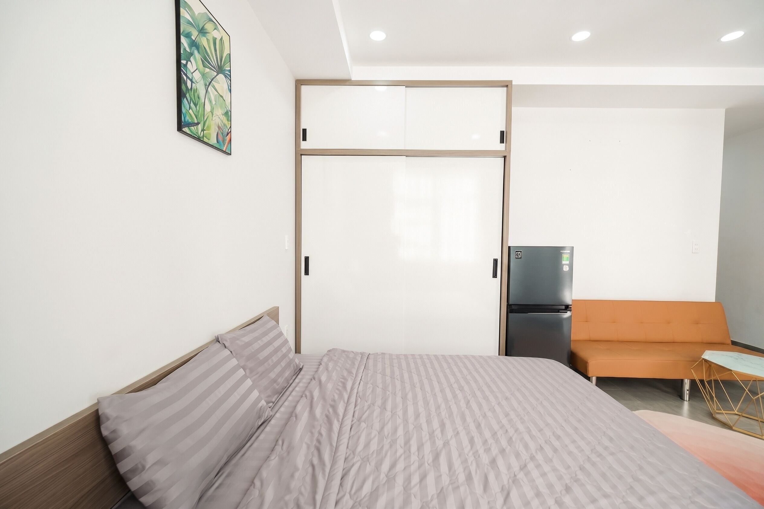 A bedroom with a bed, a sofa, a small table, a refrigerator, and a sliding door closet.