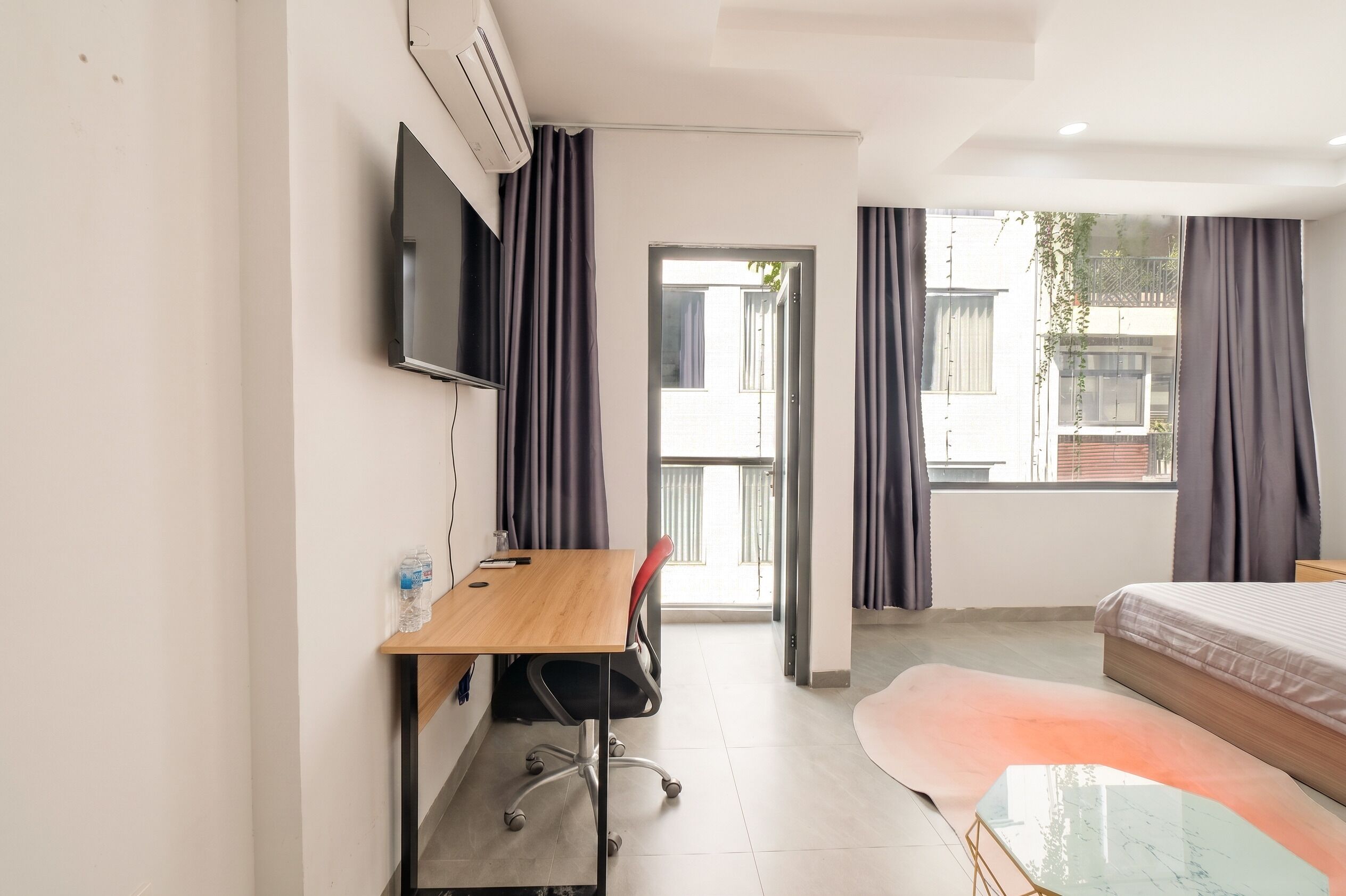 A modern hotel room with a bed, a desk with a chair, a television, and a large window with curtains.