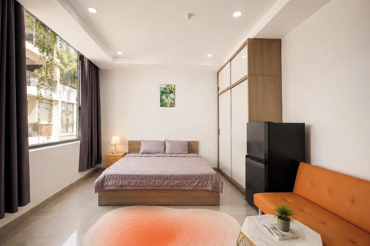 A bedroom with a bed, a sofa, a refrigerator, a nightstand, and a window with curtains.