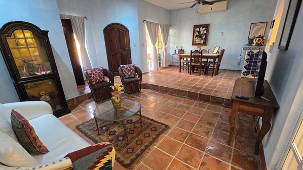 A living room with a sofa, armchair, glass coffee table, and a rug. There is a dining area with a table and chairs, a ceiling fan, and a TV stand.