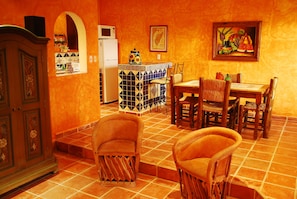 Uma sala de jantar com piso de azulejos, cadeiras de madeira, uma mesa e uma cozinha com geladeira e painel de azulejos.