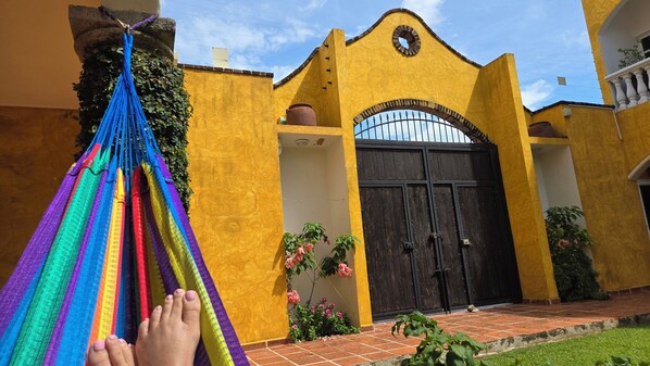 A yellow building with a large black gate, a hammock, and some pink flowers.