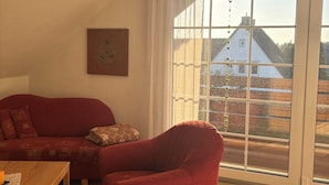 A living room with a red sofa, a wooden coffee table, and a large window with a view of a house and trees. - Beautiful top floor apartment with balcony in Osloß (Osloß)