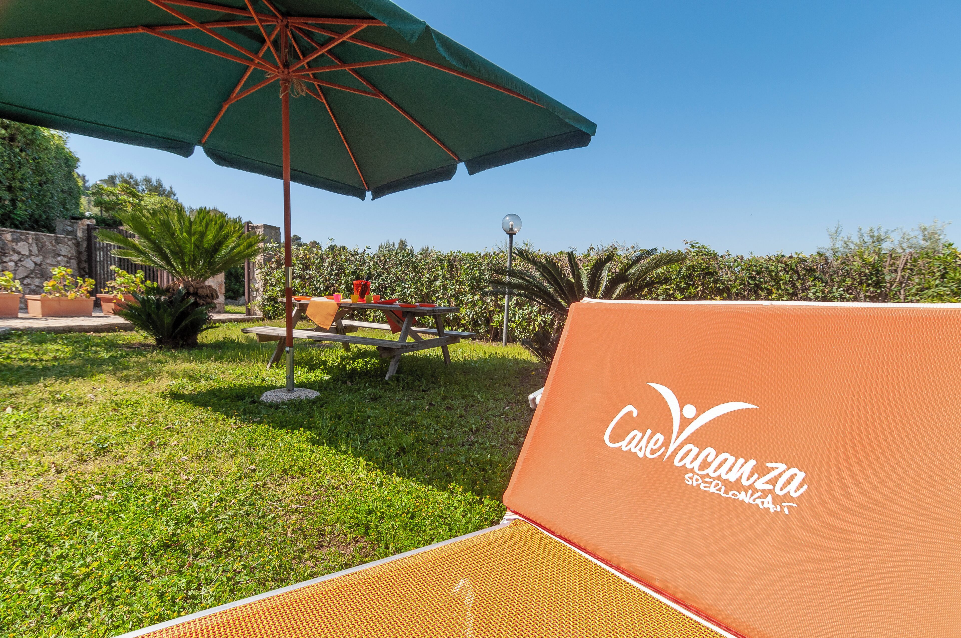 Un espacio al aire libre iluminado por el sol con una sombrilla verde, una mesa de picnic y una silla de lounge con el logotipo de "Core Vacanza".