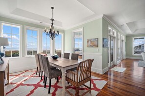 A dining room with a large table, wicker chairs, a chandelier, and large windows offering a view. - Ocean Views, Beach Right Across the Street - 714 Winter Trout (Fripp Island)