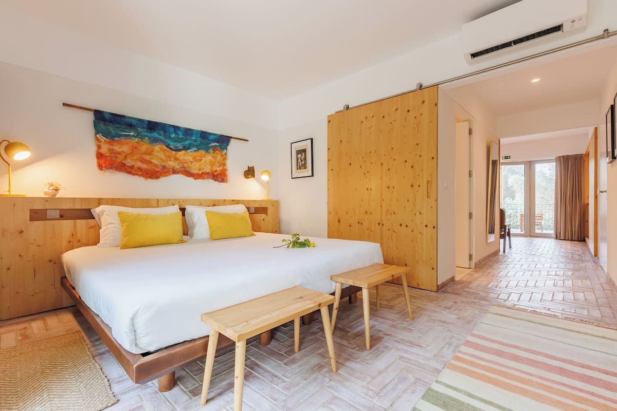 A modern hotel room with a large bed, wooden headboard, bedside lamps, a colorful abstract painting, and a view of the hallway.
