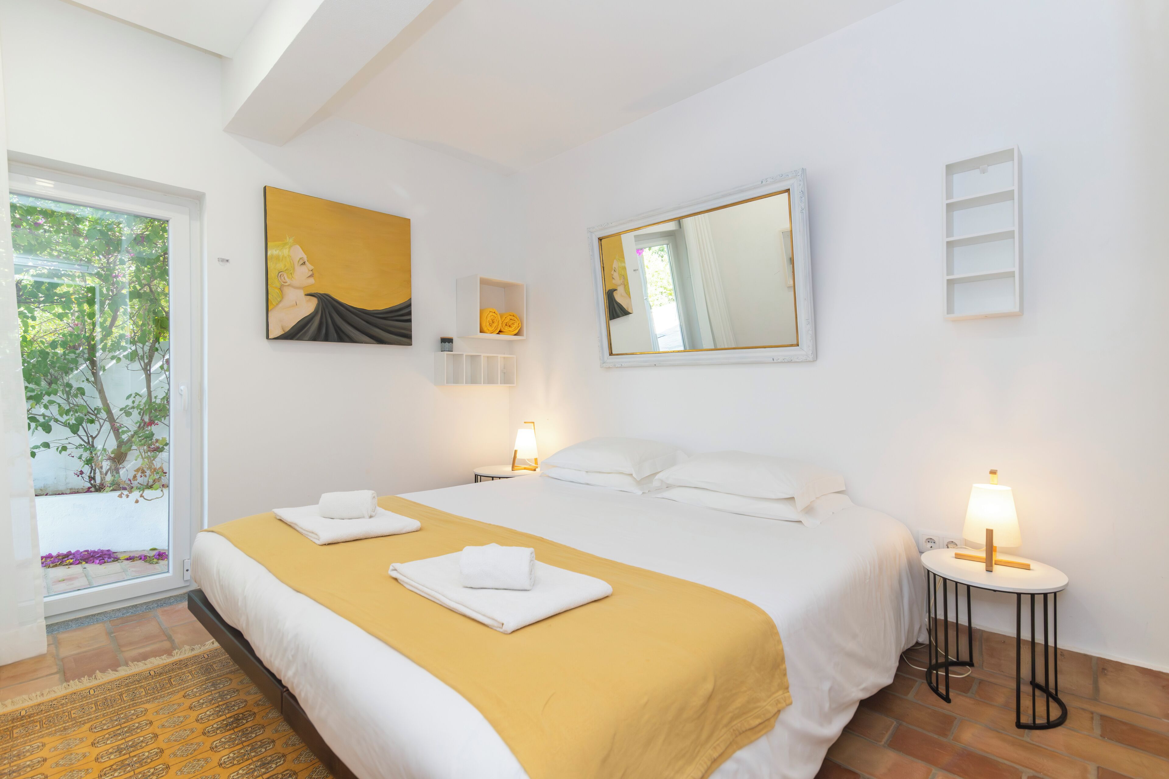 Habitación de hotel con cama, dos mesitas de noche con lámparas, un espejo grande, un cuadro y una ventana con vistas a árboles.
