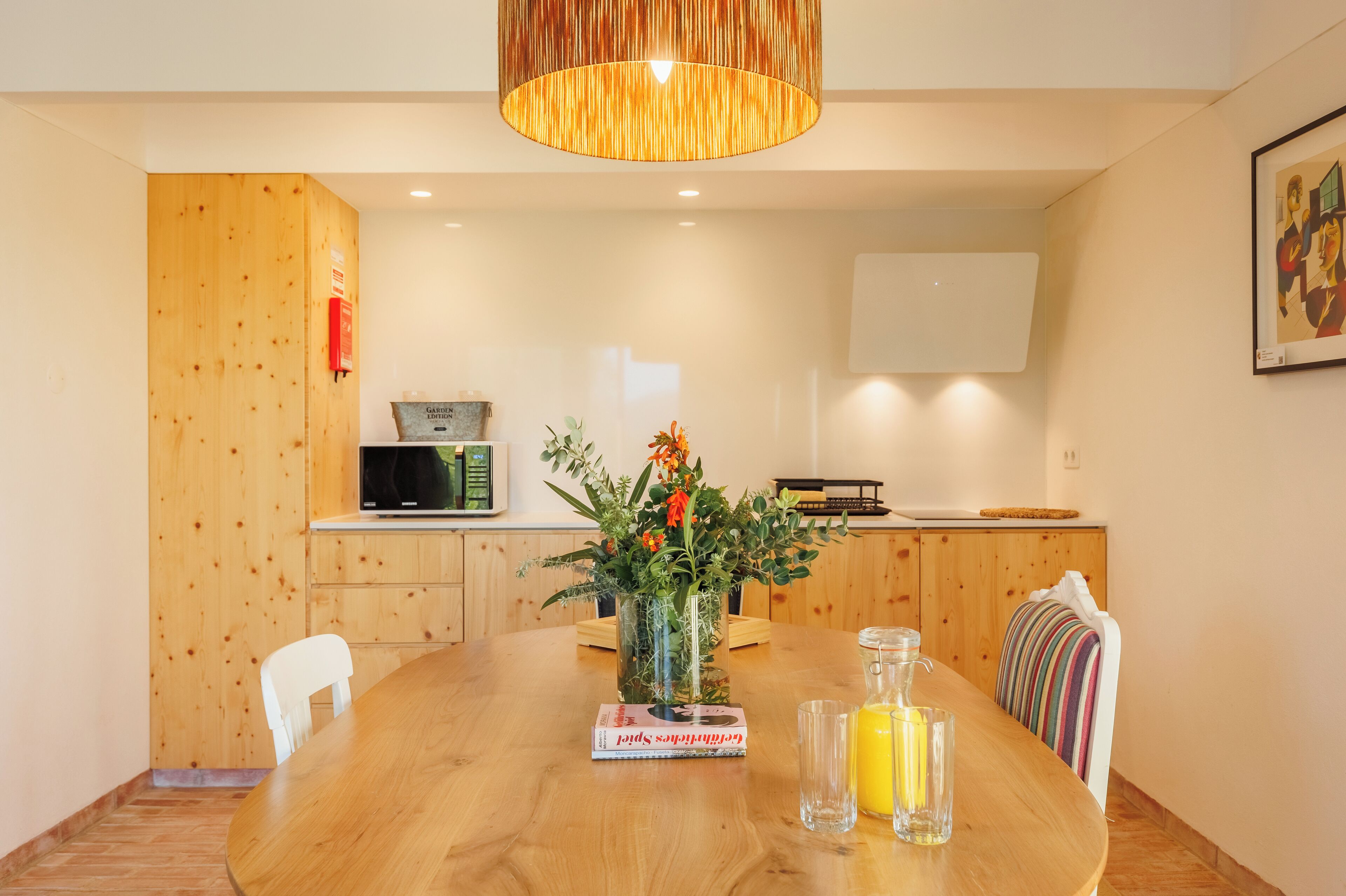 A dining area with a wooden table, chairs, a vase with flowers, a book, and a glass bottle.