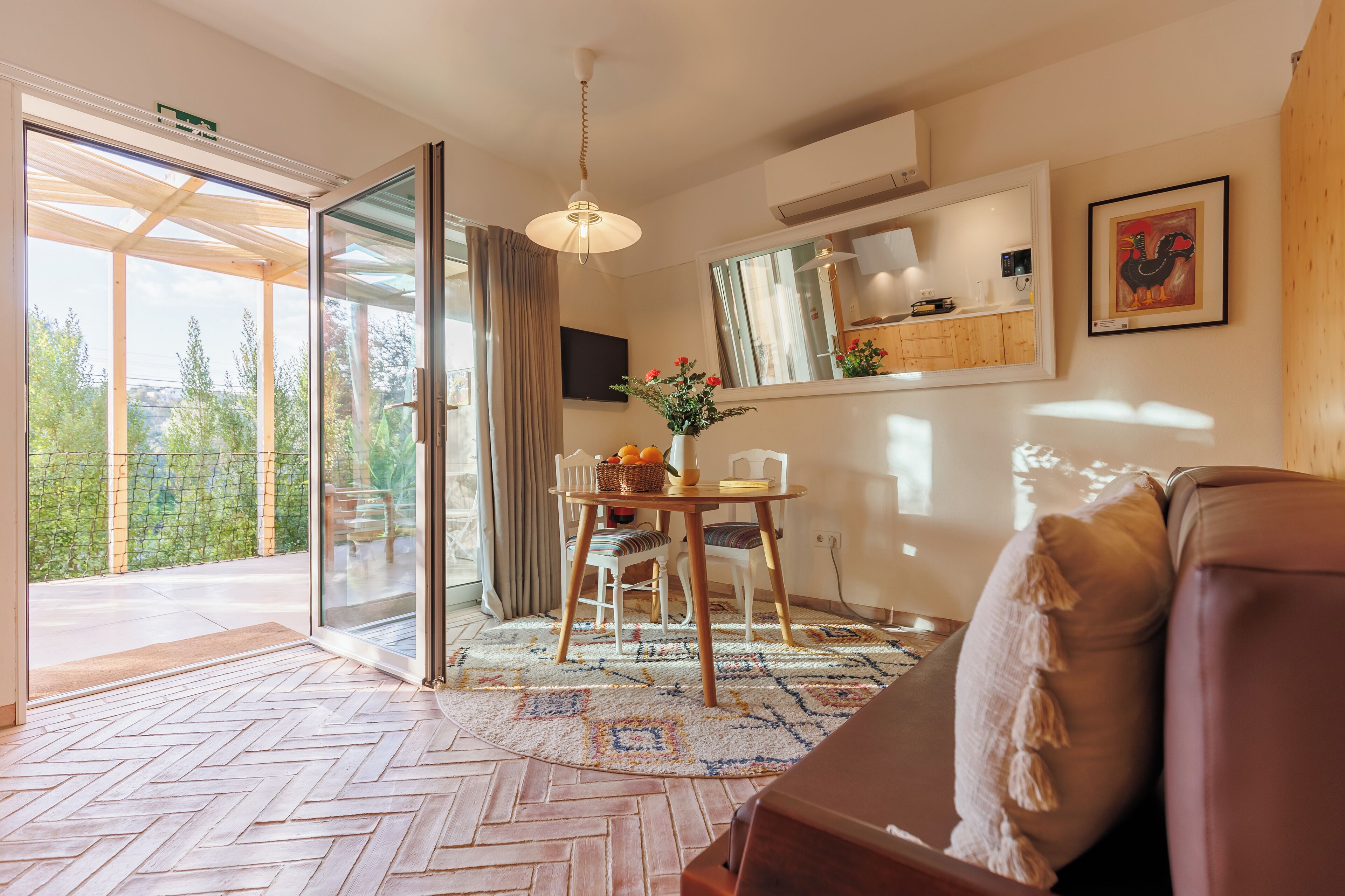 Una sala moderna con suelo de espiga, un sofá de cuero marrón, un comedor con mesa y sillas, un ventanal con vistas a la vegetación y un ventilador de techo.