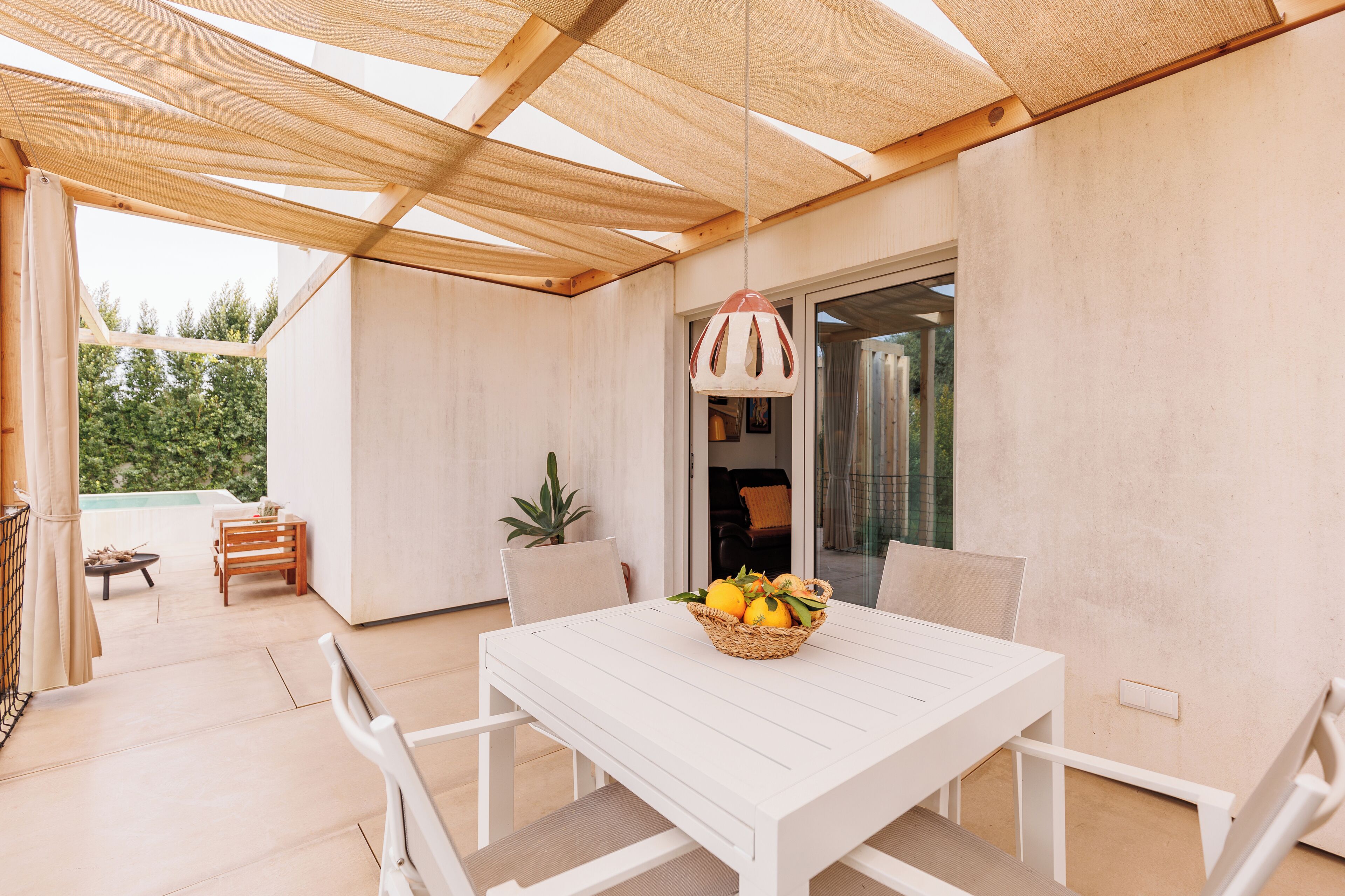 A white table with chairs and a basket of fruit in a sunlit room with a wooden ceiling and a view of greenery outside.