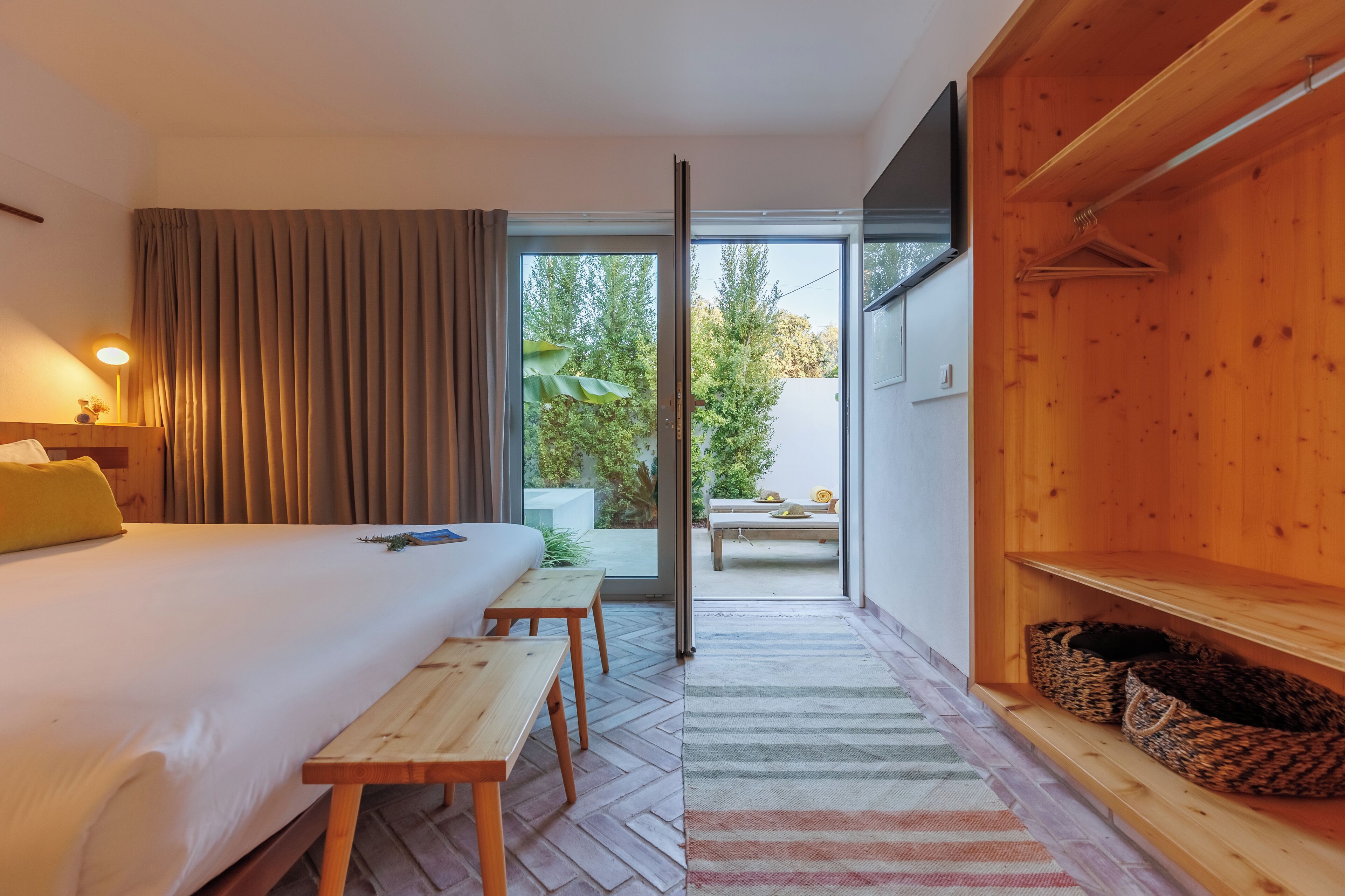 Quarto de hotel com cama, um banco de madeira, uma mesa, TV, porta de vidro deslizante para área externa com mesa e cadeiras e prateleira de madeira com cestos.