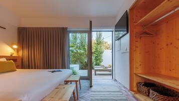 Quarto de hotel com cama, um banco de madeira, uma mesa, TV, porta de vidro deslizante para área externa com mesa e cadeiras e prateleira de madeira com cestos.