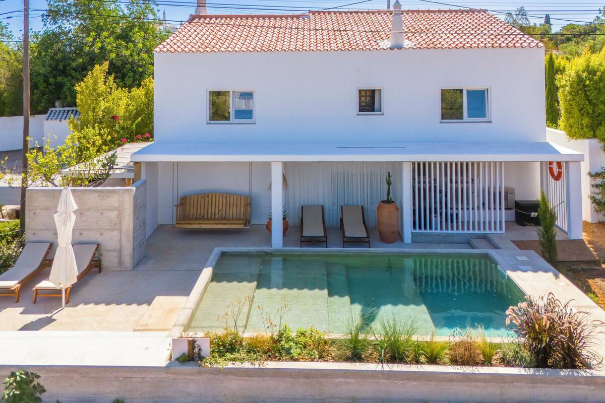 A two-story house with a swimming pool, a covered patio with lounge chairs, and a small garden area.