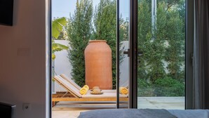 A room with a large window, a bed, and a view of a patio with a lounge chair, a large pot, and greenery. - Laranjal Farm House (Olhao)