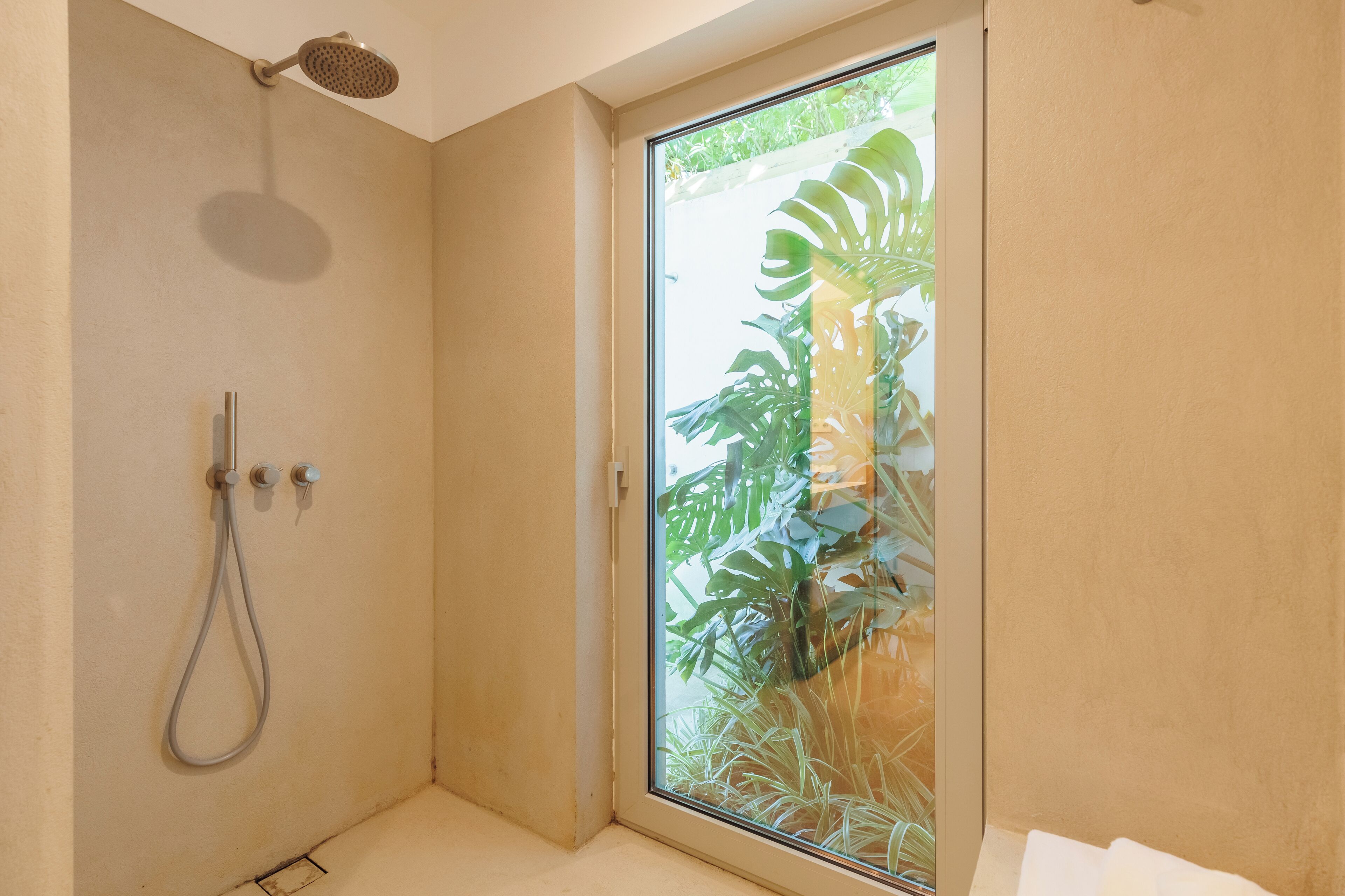 A shower with a handheld showerhead and a handheld hose, a sliding glass door leading to a garden with large tropical plants.
