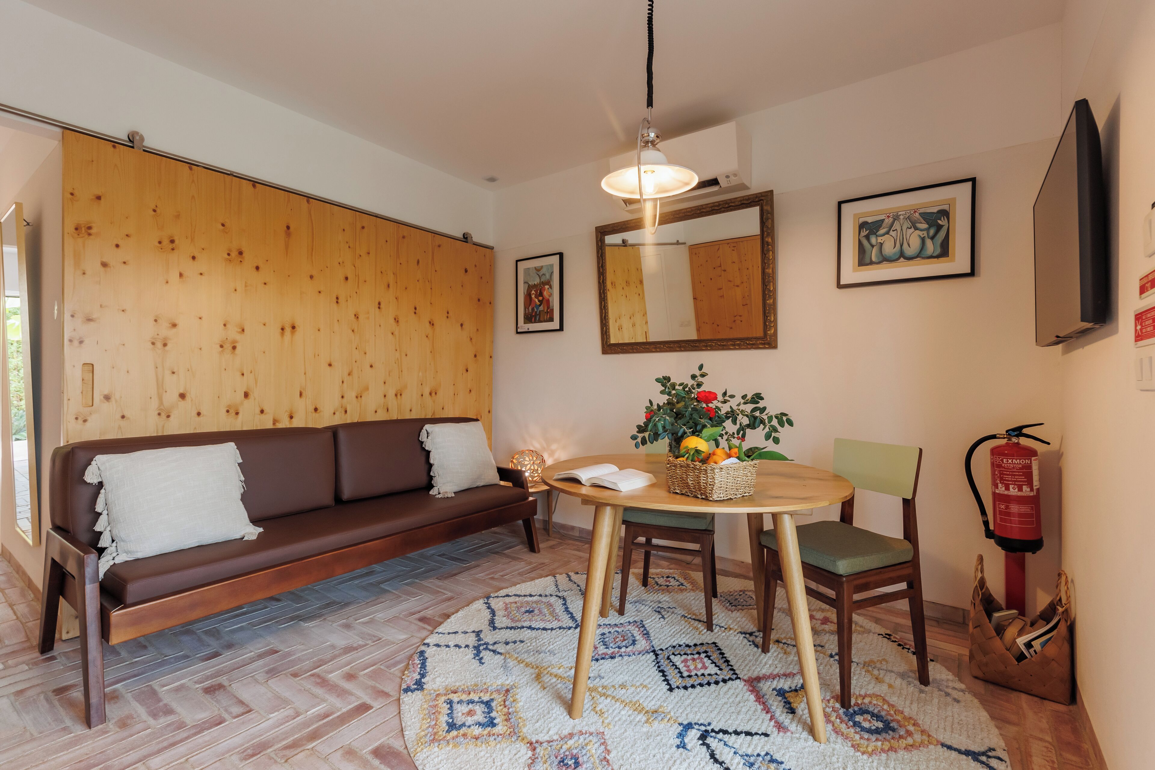 A cozy living room with a brown sofa, a wooden wall, a round table with a basket of fruit, and a fire extinguisher.