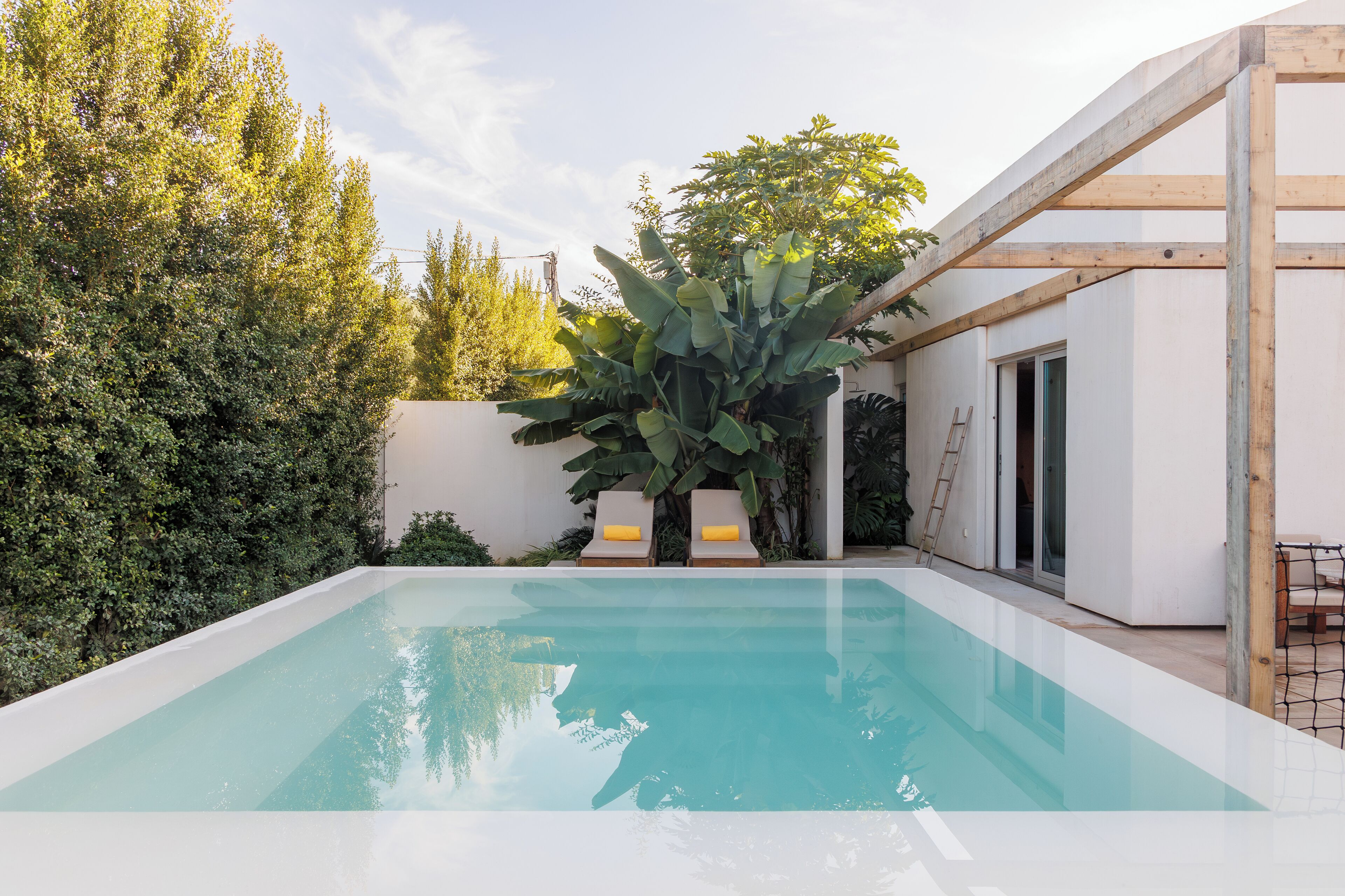 A rectangular swimming pool with a clear blue water surface, surrounded by lush greenery and a white building with a wooden structure on one side.