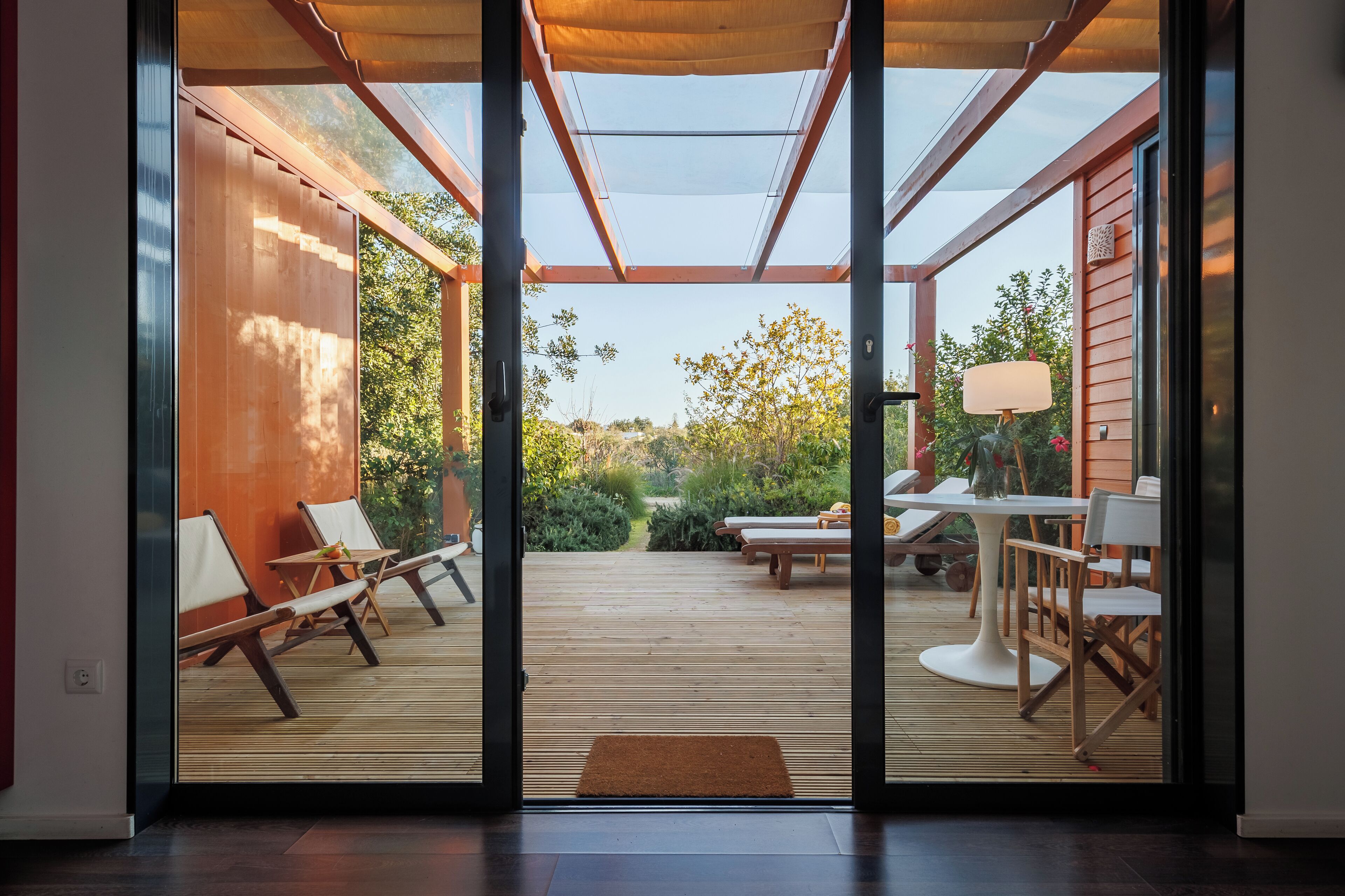 Un patio moderno con suelo de madera, mobiliario exterior y una puerta de cristal que da a un jardín.