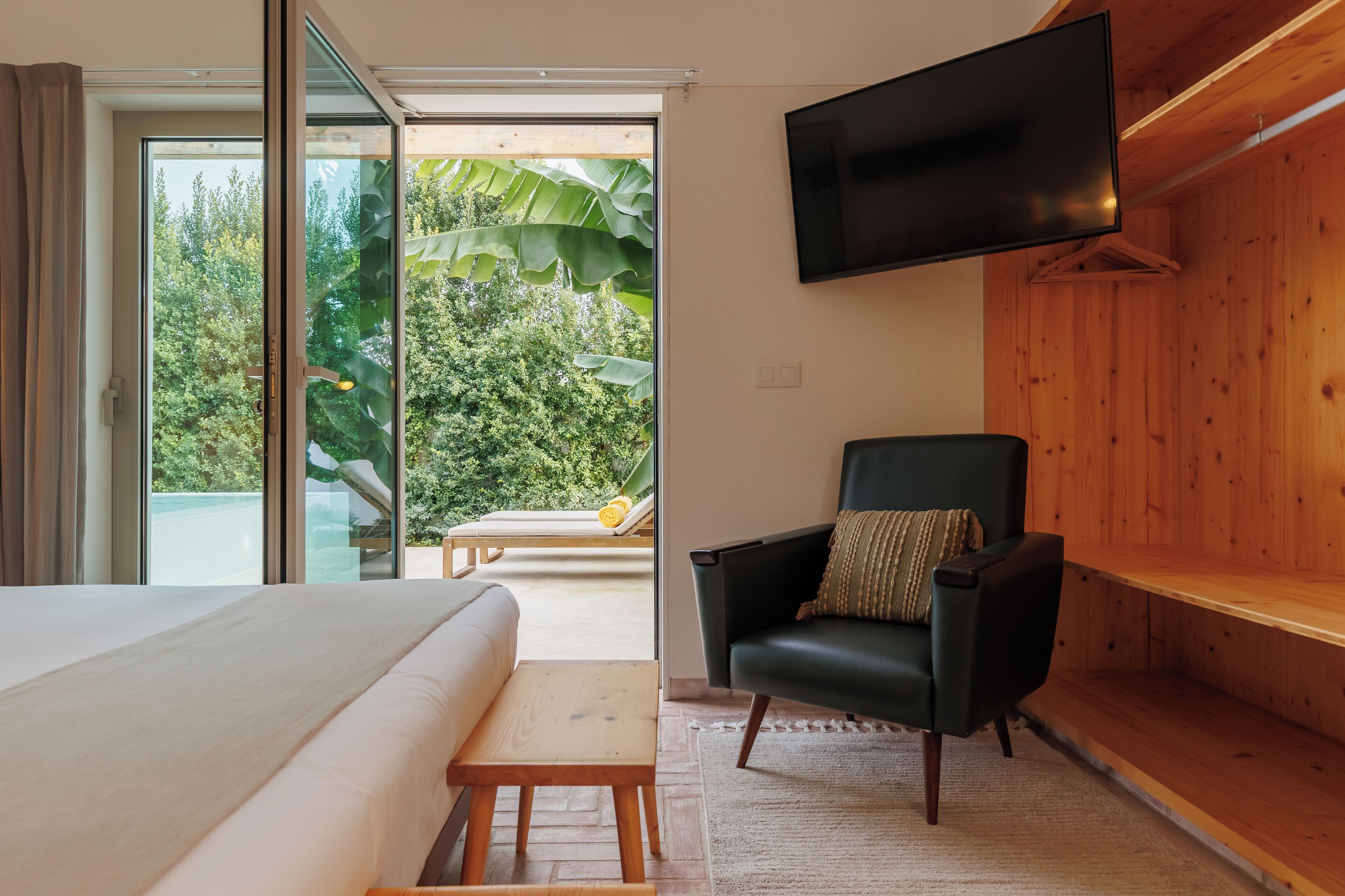 A bedroom with a bed, a chair, a TV, and a view of a pool and greenery.