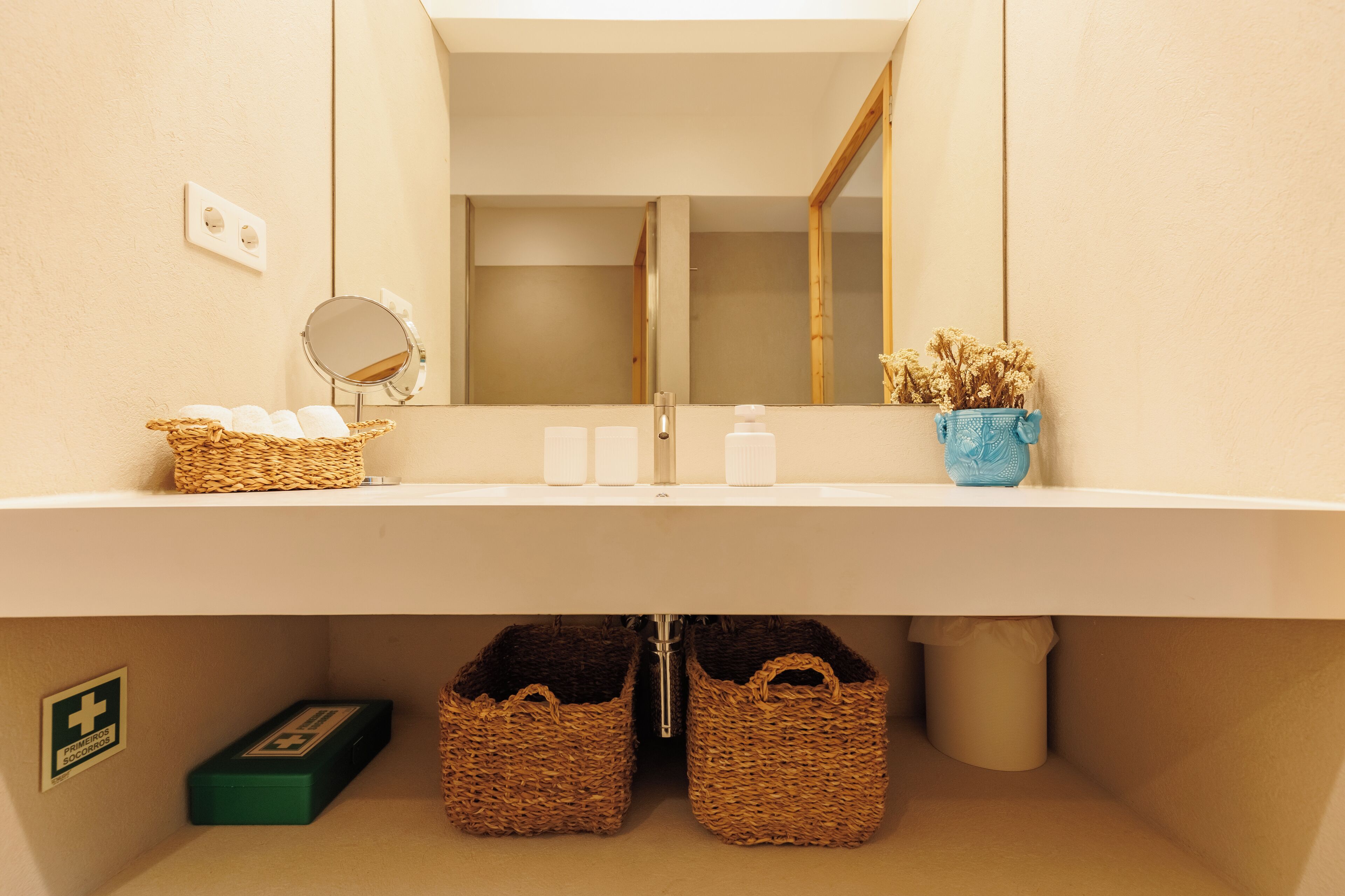 A bathroom with a white sink, a mirror, a basket of toiletries, and a vase with dried flowers.