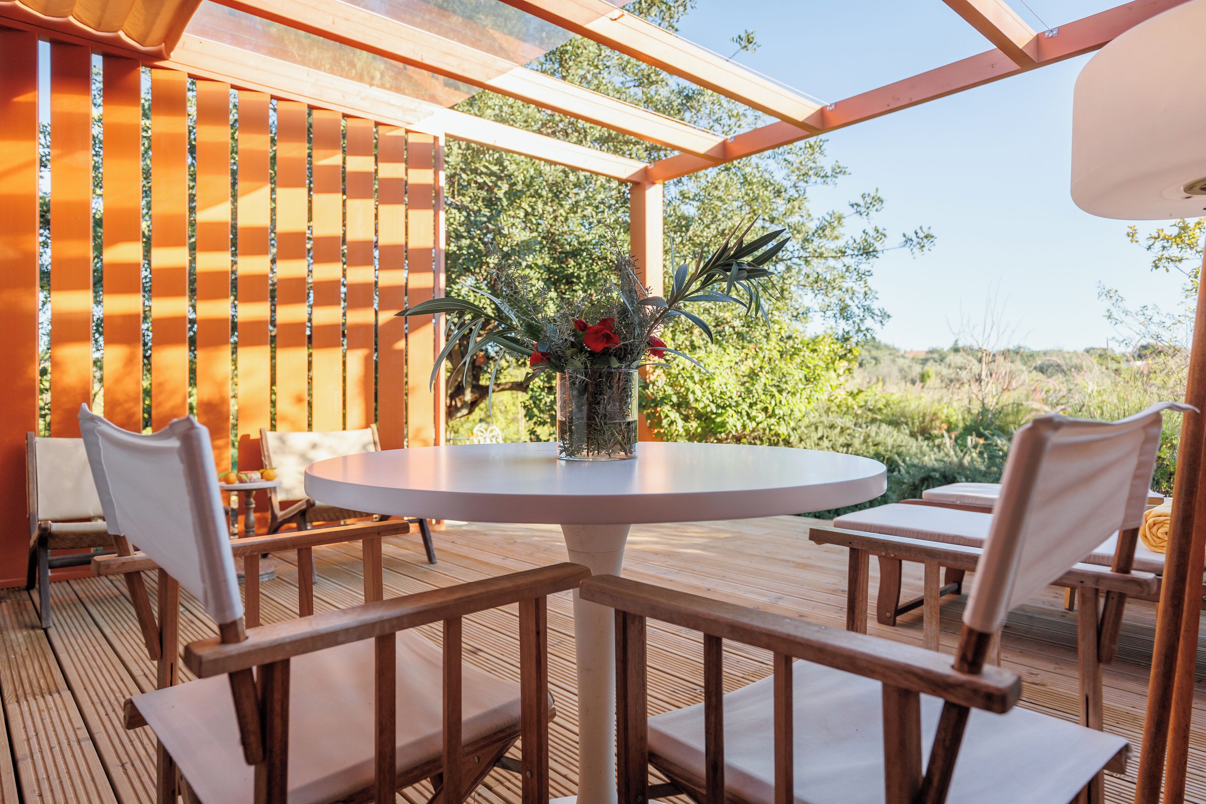 Een houten terras met een ronde tafel en stoelen, een glazen overkapping en uitzicht op groen.