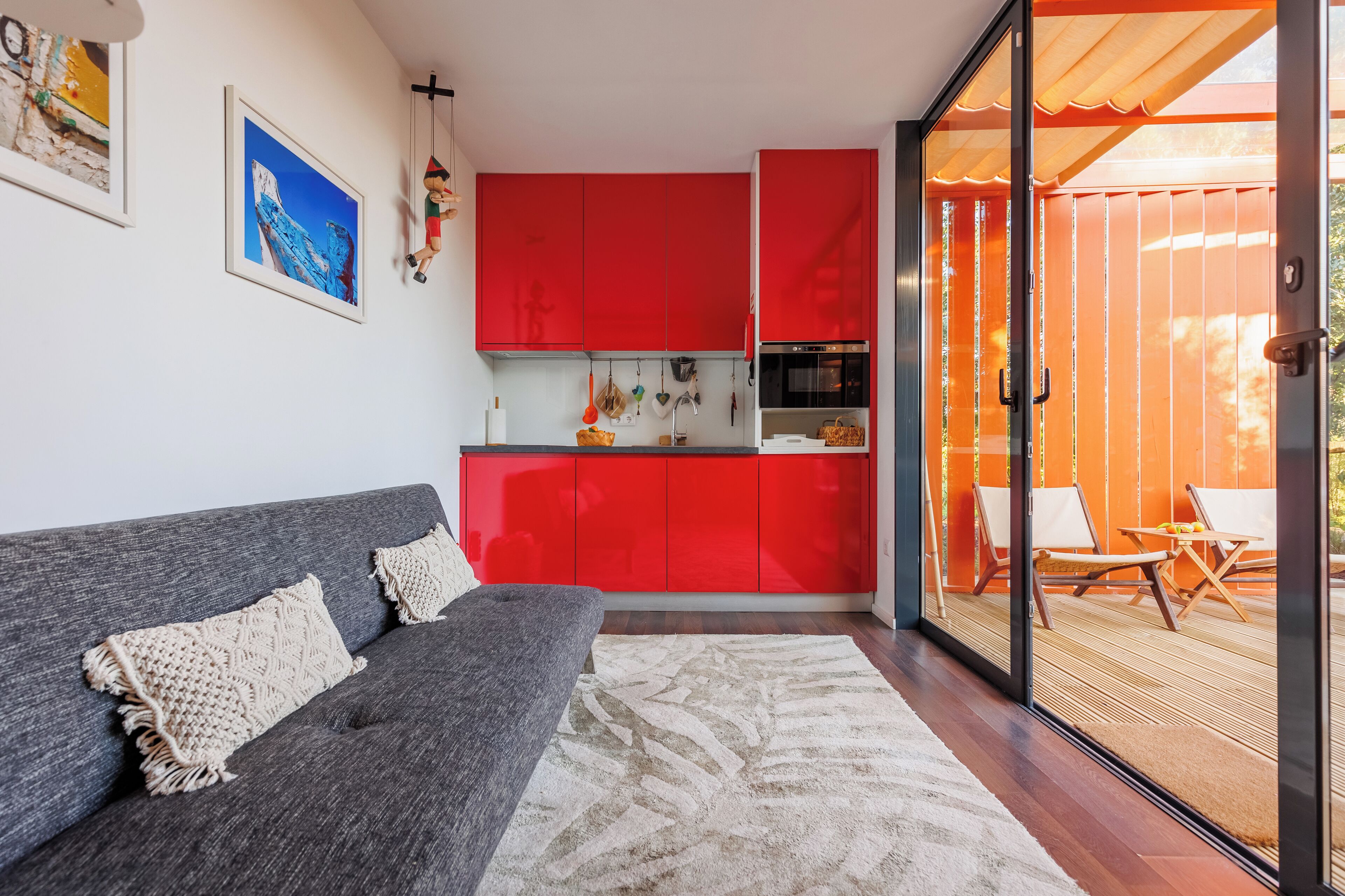 Una sala moderna con un sofá gris, un ventanal grande y una cocina roja al fondo.